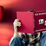 A man is reading a red book in front of a red wall and next to a red lamp.