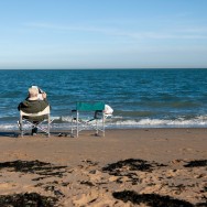 An elderly man is sitting in a seat at the beach while reading the newspaper, there is an empty seat next to him.