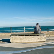 A man sits near a beach and watches ships go sailing by.