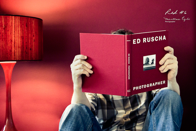 A man is reading a red book in front of a red wall and next to a red lamp.