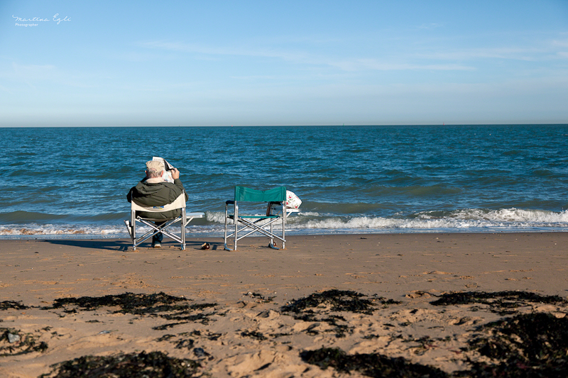 An elderly man is sitting in a seat at the beach while reading the newspaper, there is an empty seat next to him.